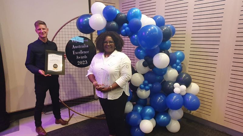 Nicholas Hill and Bosede Adetifa holding a glass trophy and awards certificate in front of a blue and white balloon garland. Nicholas Hill and Bosede Adetifa holding a glass trophy and awards certificate in front of a blue and white balloon garland. Nicholas Hill and Bosede Adetifa holding a glass trophy and awards certificate in front of a blue and white balloon garland.
