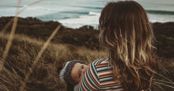 A woman is standing on a hill looking at the ocean. She has her back to the camera and she appears to be breastfeeding a small baby.
