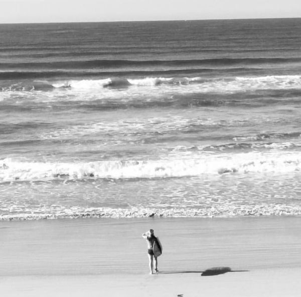 A woman stands on the shore alone holding her suf board. There is no one else around and the ocean is large and looming.