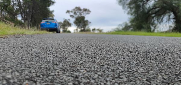 View down a road from ground level with a blue car parked ahead on the side of the road. Trees and grass are either side