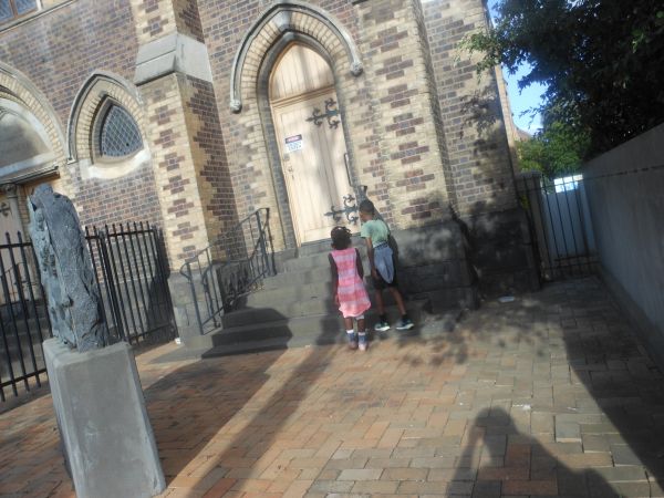 The image shows an entrance to a church, two children a boy and a girl walking towards the wooden door. There is a shadow of a person taking the photograph behind them.
