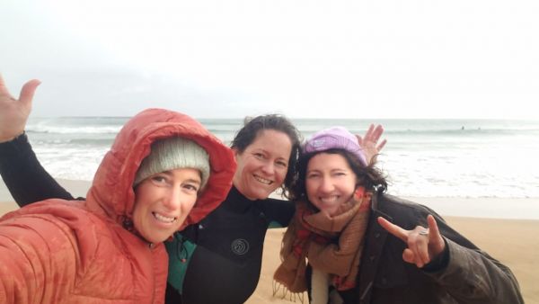 Three women standing on the sand in front of the ocean wearing winter clothes. All the women are smiling