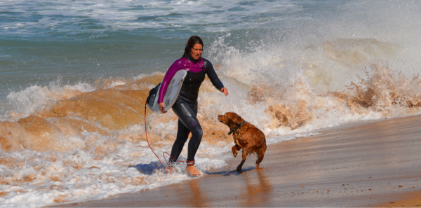 A woman is walking out of the ocean with a wave crashing behind her. She is holding a surfboard and her dog is running towards her on the shore.