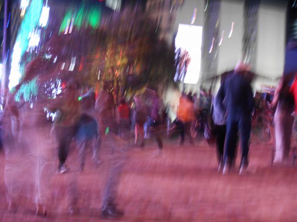 The image is blurry and has a dreamlike quality, suggesting a busy, bustling scene. The colors are vibrant, and the atmosphere is festive. There are many people moving around, creating a sense of motion. The people are blurred and indistinct, but their shapes and colors are visible.