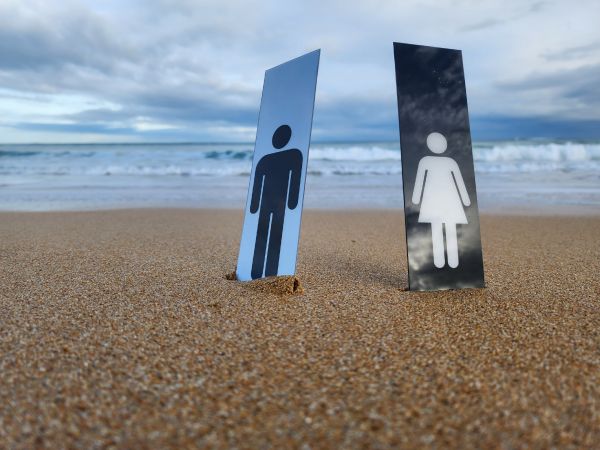 Two signs are wedged into the sand in front of the ocean. One is white and is of a male icon, the other is black and is of a woman icon.