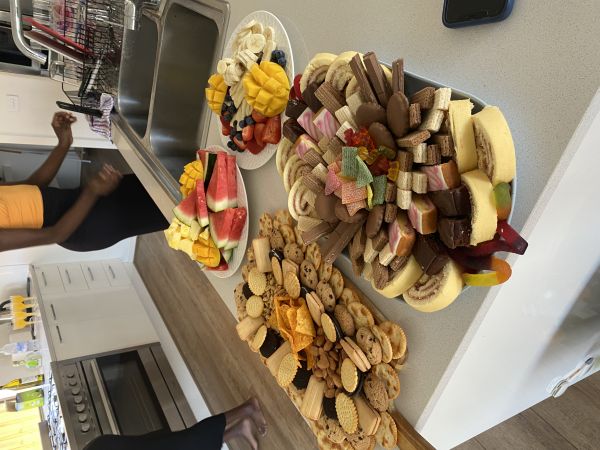 A kitchen bench with platters of biscuits, cakes, and fruit, with two people standing in the background.