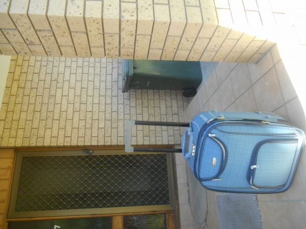 A blue, greyish suitcase with wheels standing on a porch outside in front of a brick house.