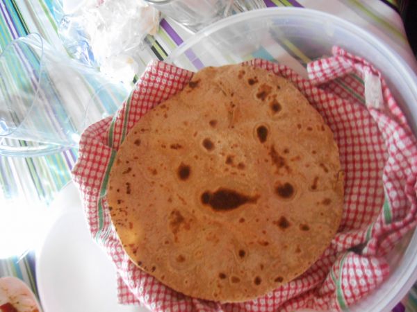 A roti, round, flatbread resting on a red and white and red cloth inside a clear plastic container. The container is sitting on a table with a colorful tablecloth.