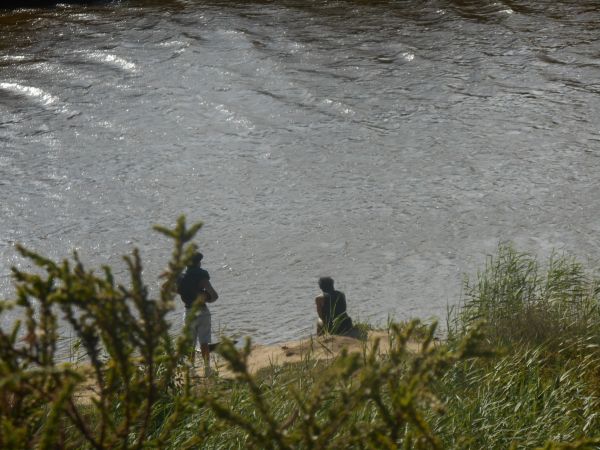 A vast green landscape by the river. Two silhouettes are placed in the middle, showing two young people sitting by the river and enjoying the view.