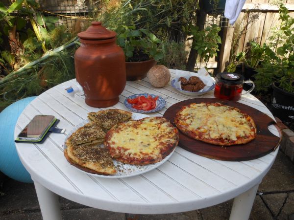 The image shows a table set up outdoors for a meal. There are various dishes of food on the table, along with drinks and other items. The setting is a garden with lush greenery.