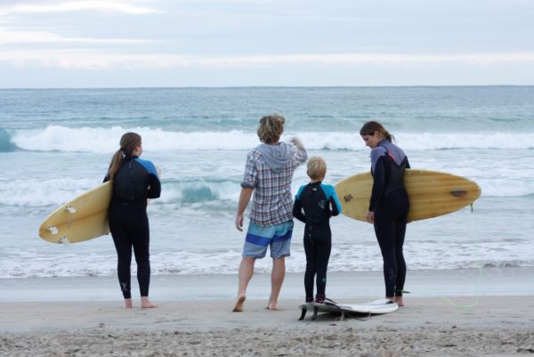A mother, father and two children standing on the sand with their backs to the camera. The mother and two children have surf boards and are wearing wetsuits. The father is in normal clothes.
