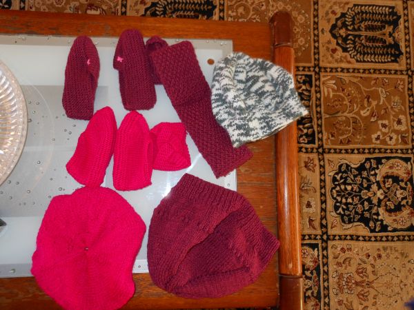 A variety of knitted items laid out on a table. The are in various shades of red and grey. There is a beret, scarf and mittens.