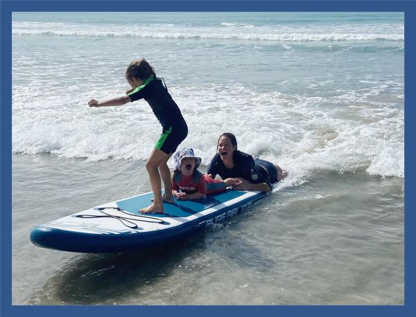 A woman and 2 young girls are on a long surfboard catching a small wave in the shallow ocean. Everyone is smiling.