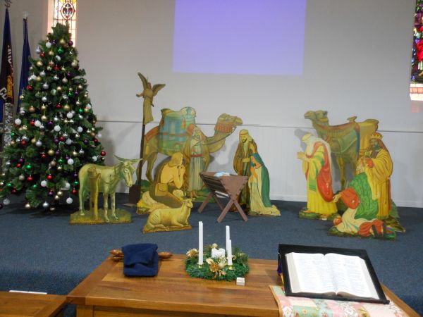 The image shows a nativity scene set up in a church. There is a Christmas tree decorated with ornaments, a table with candles and a Bible.