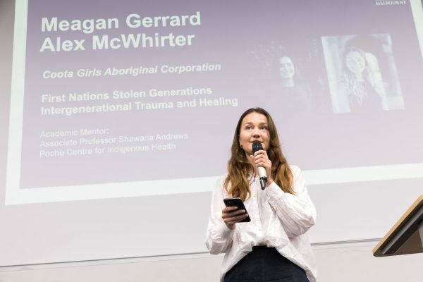 Alex McWhirter speaking into a microphone. A slide behind her shows her name, fellows project and mentor