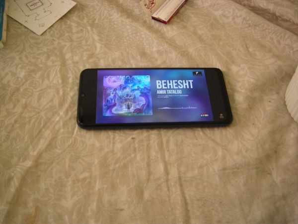  The image likely depicts someone listening to music on their smartphone. The phone is on the bed. The screen of the phone is lit up, displaying a music player app. The song title that appears is 