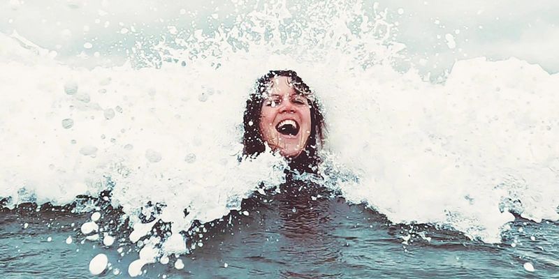 A women's face emerging from a wave. Her mouth is open and she has a large smile.